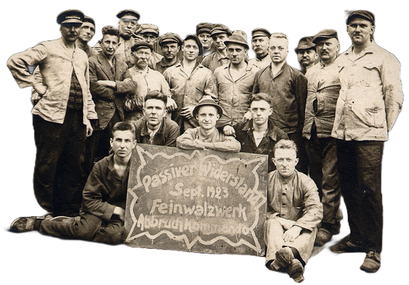 Das Bild ist eine Schwarz-Weiß-Aufnahme aus 1923, die eine Gruppe von etwa zwanzig Männern zeigt, die in Arbeitskleidung und oft mit Mützen oder Kappen bekleidet sind. Einige der Männer sitzen oder knien im Vordergrund, während die Mehrheit dahinter steht. In der Mitte des Vordergrunds halten drei Männer ein Schild hoch. Auf dem Schild steht in geschwungener Schrift: "Passiver Widerstand Sept. 1923 Feinwalzwerk Abbruch Kommando". Die Männer wirken ernst und nachdenklich, einige blicken direkt in die Kamera.