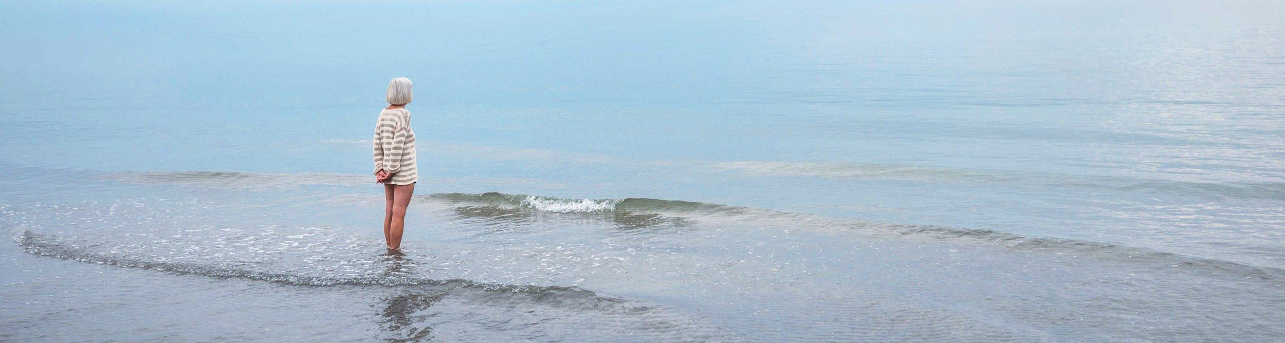 das Bild zeigt eine Person mit hellen kurzen Haaren am Strand blickt aufs ruhige Meer.